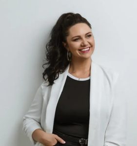 Woman with long curly dark hair in a ponytail, wearing a white blazer over a black top, smiling and looking to the side against a plain white background.